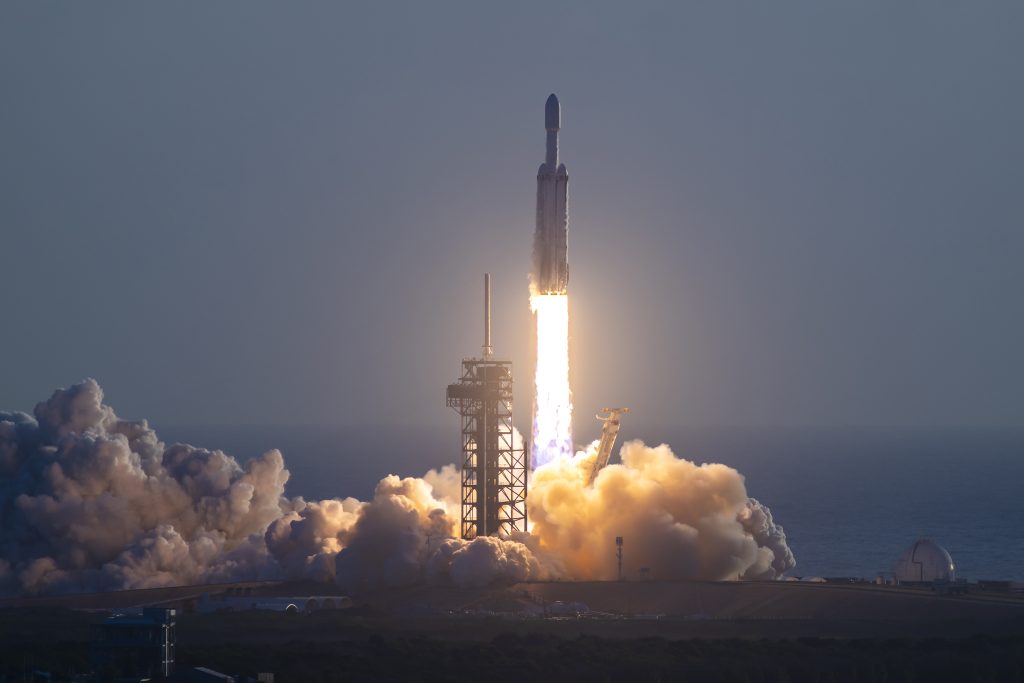 A rocket lifts off from a coastal launch pad, sending bright flames and a plume of white smoke across the ground.